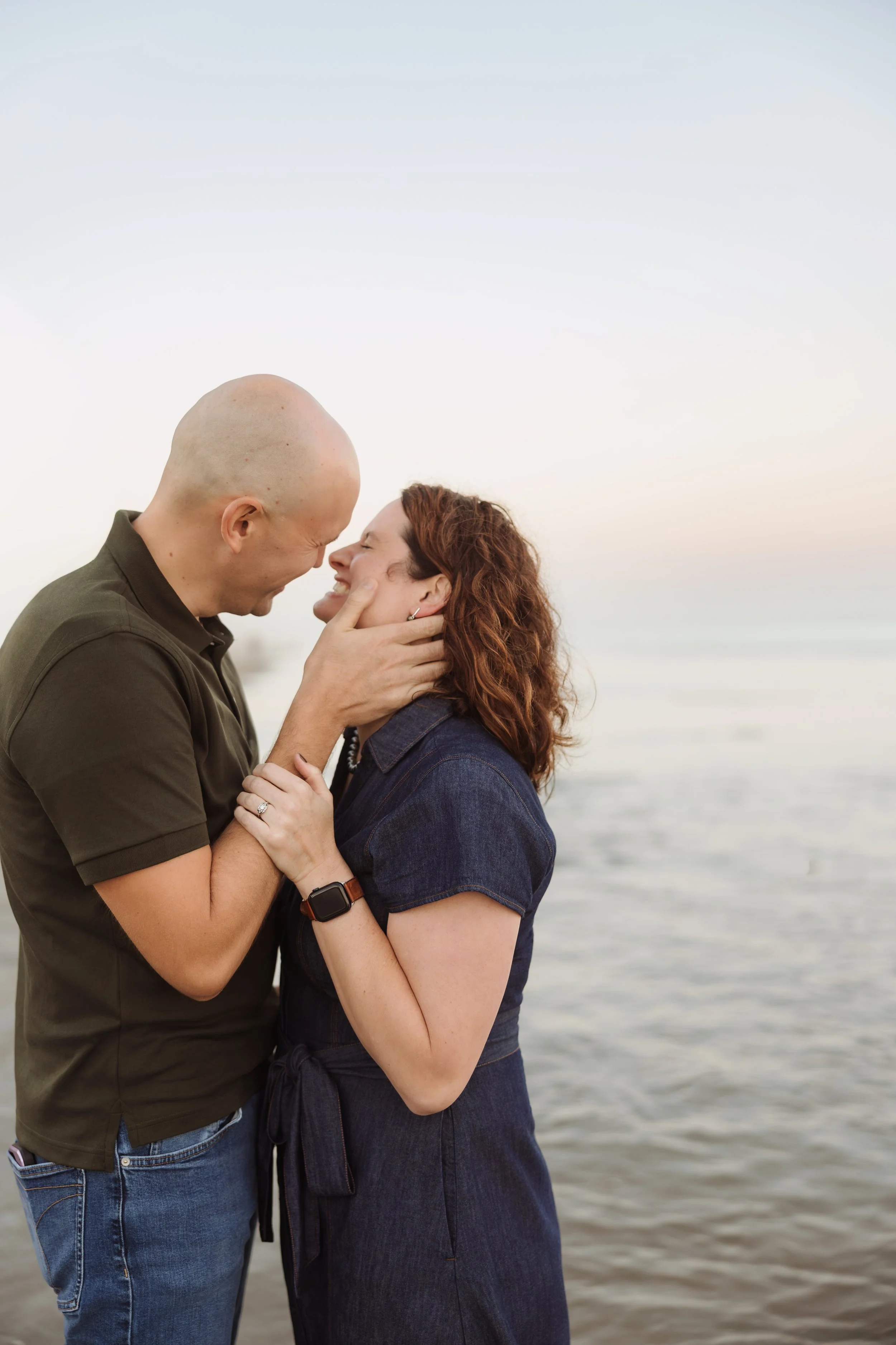 A couple is standing close together near the water at sunset, smiling and touching foreheads, with the man gently holding the woman's face.