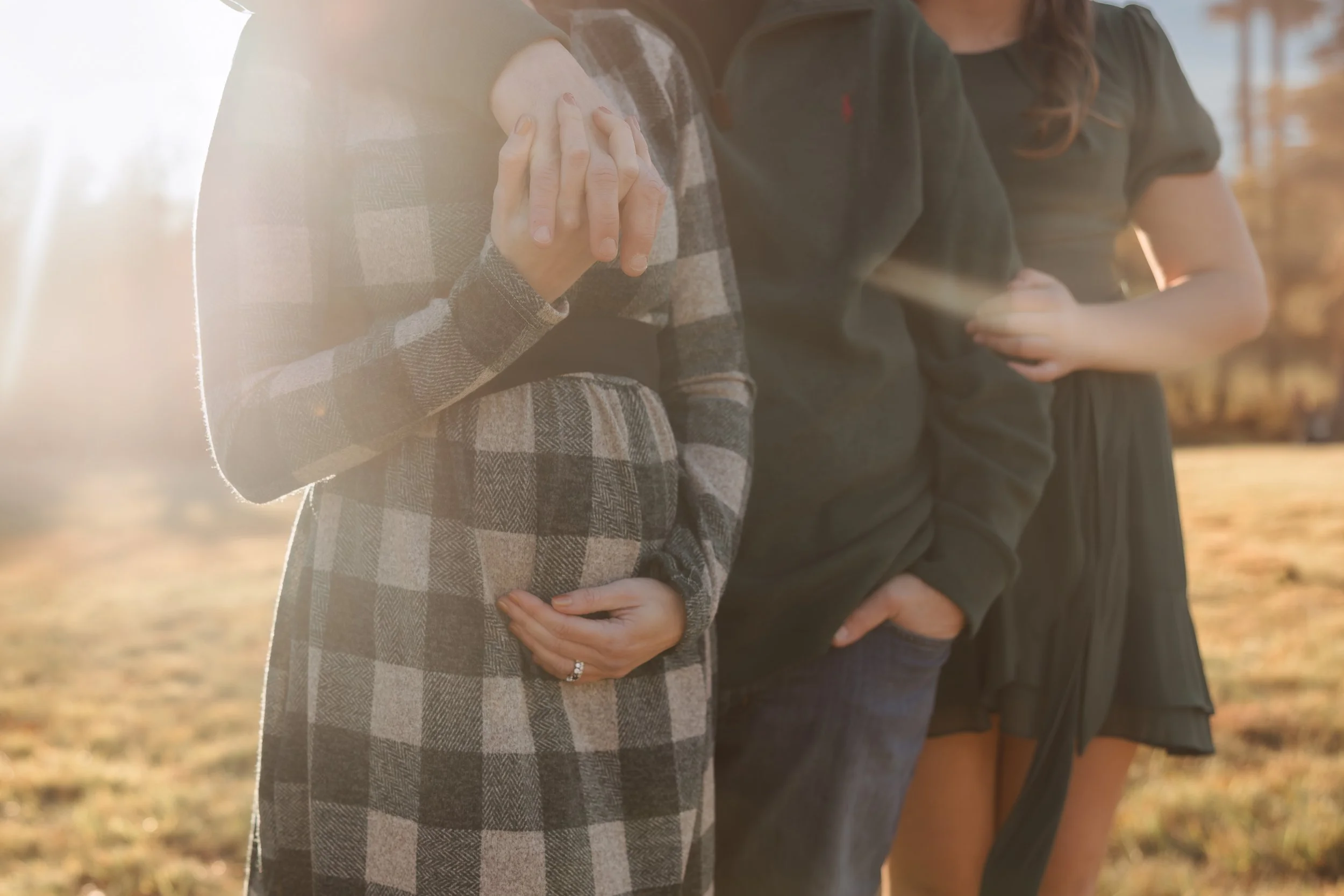 Three women standing outdoors during sunset, with two women holding hands and one resting her hand on her stomach, all dressed in casual clothing.
