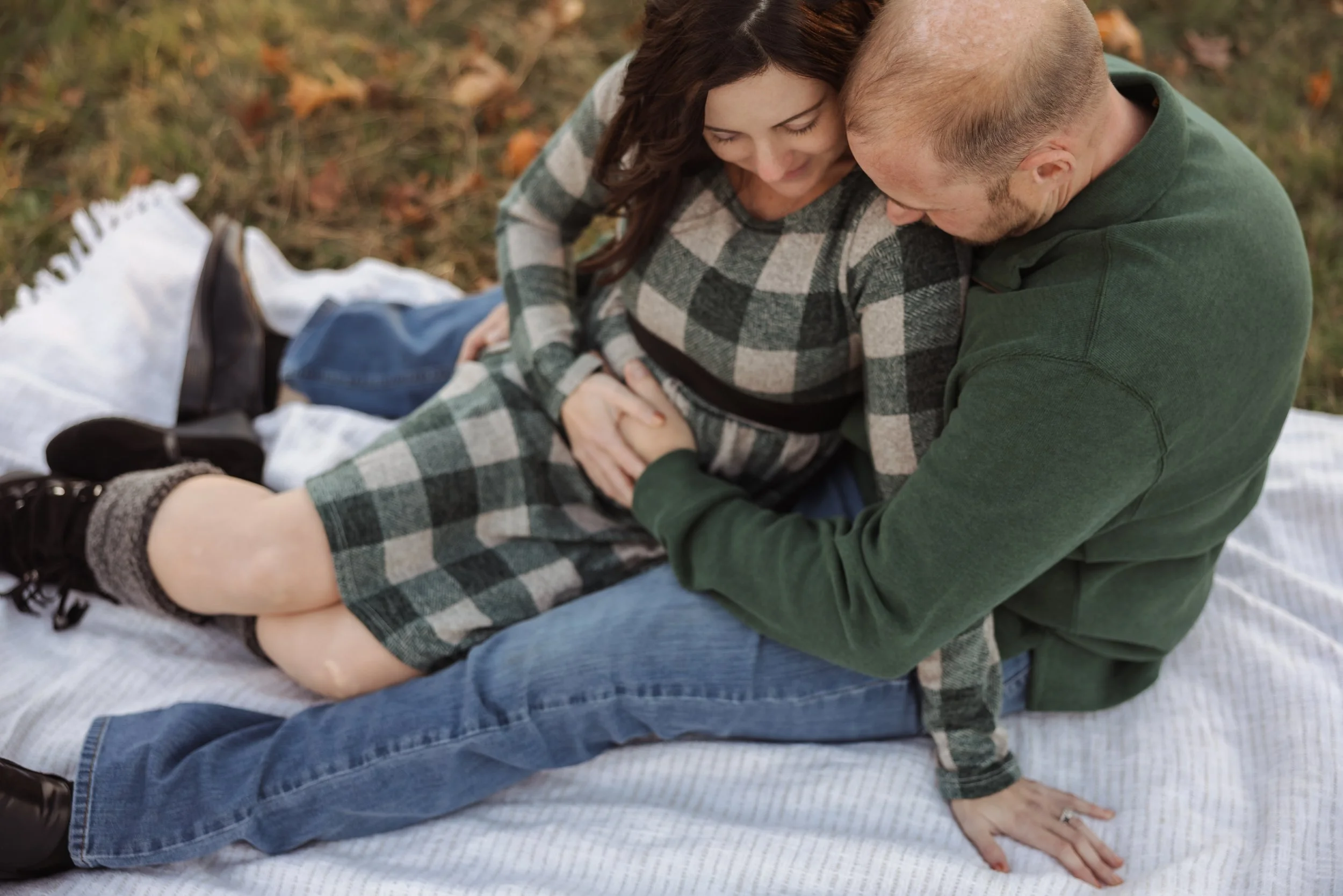 A couple sitting on a blanket outdoors, with the woman leaning on the man, both smiling and close together in a forested area with fallen leaves.