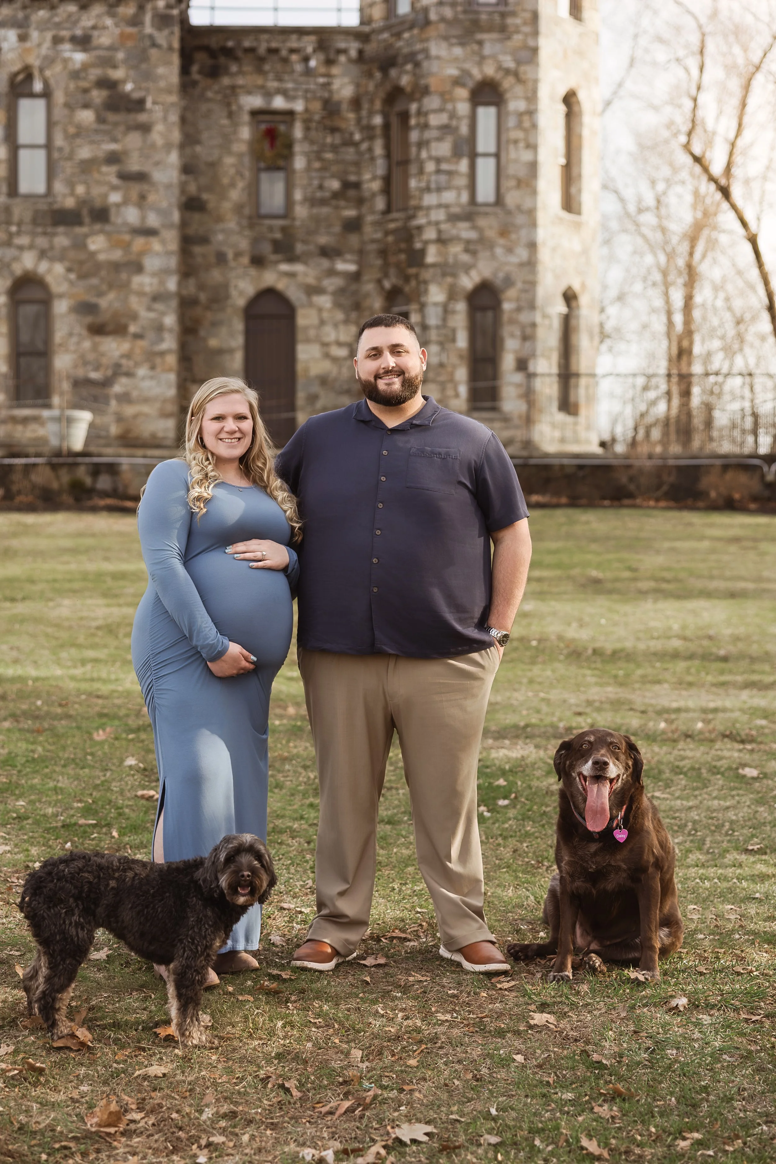 A pregnant woman in a blue dress, smiling, standing next to a smiling man in a dark shirt, with two dogs sitting on the grass in front of them, in front of a stone building.