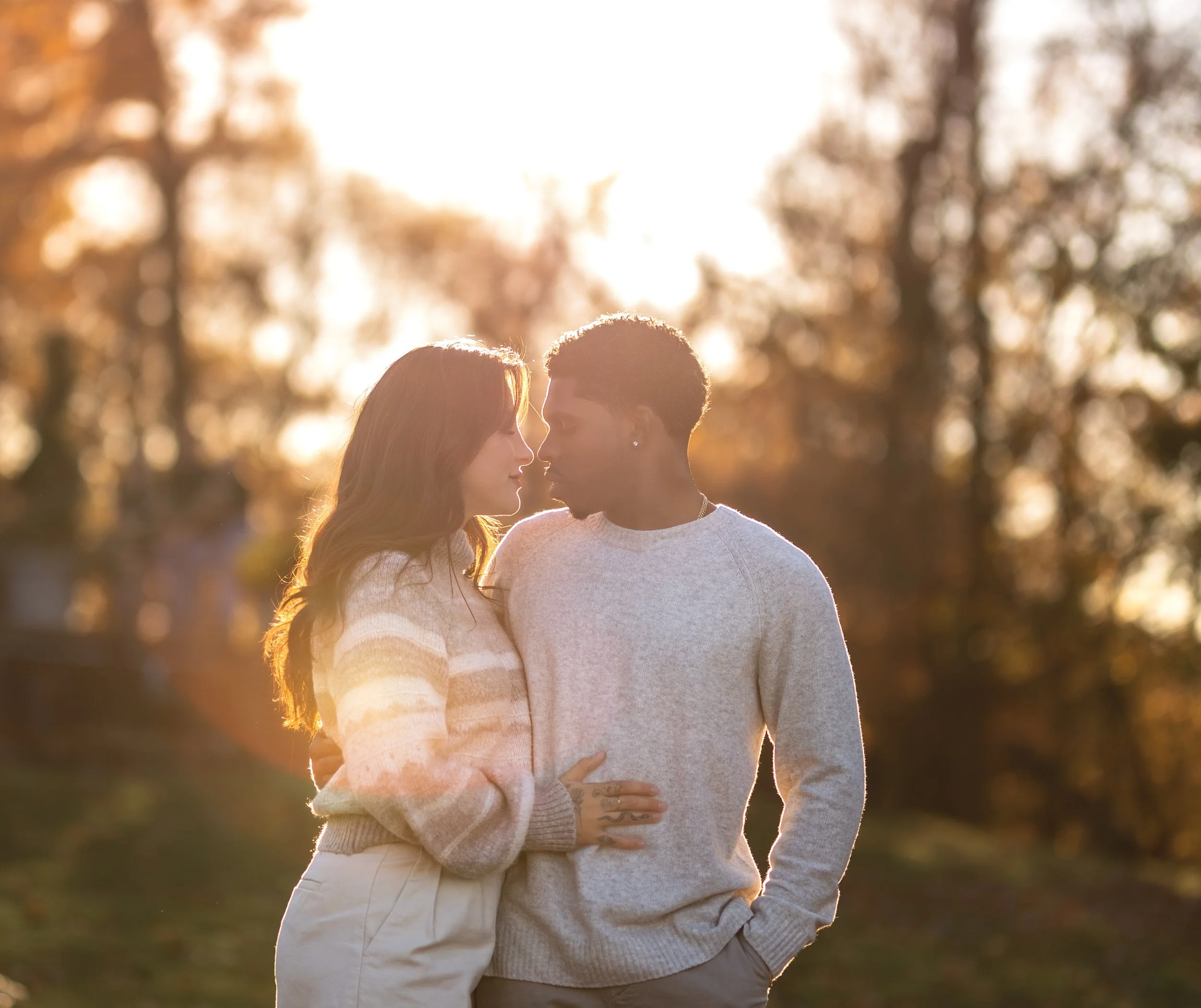 A couple stands close together outdoors during sunset, with trees blurred in the background. The woman has long hair and is wearing a light-colored sweater, and the man has short hair and earrings, wearing a light gray sweater. They are gazing at eac