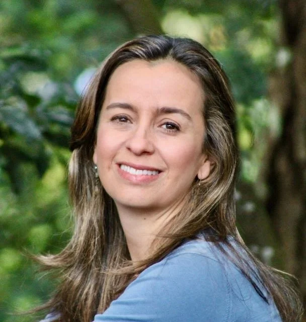 A woman smiling outdoors with green foliage in the background.