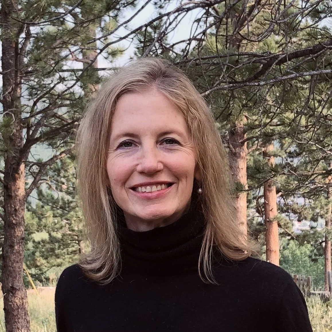 A smiling woman with shoulder-length blonde hair, wearing a black turtleneck, standing outdoors in front of trees in a forested area.