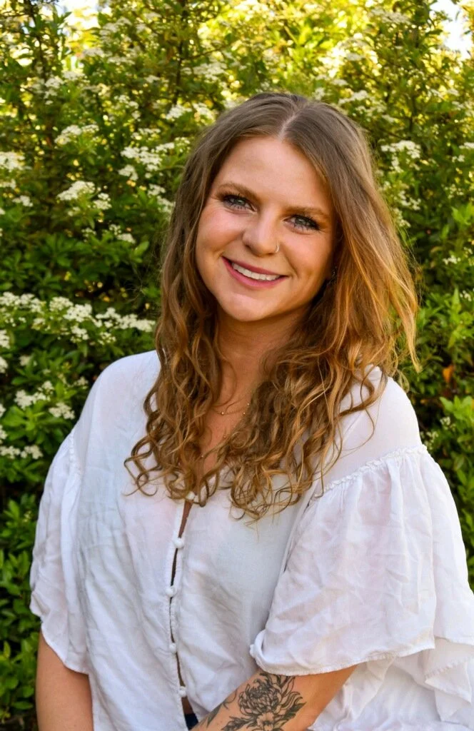 Portrait of a smiling woman with wavy light brown hair, wearing a white blouse, standing outdoors with greenery and white flowers in the background.
