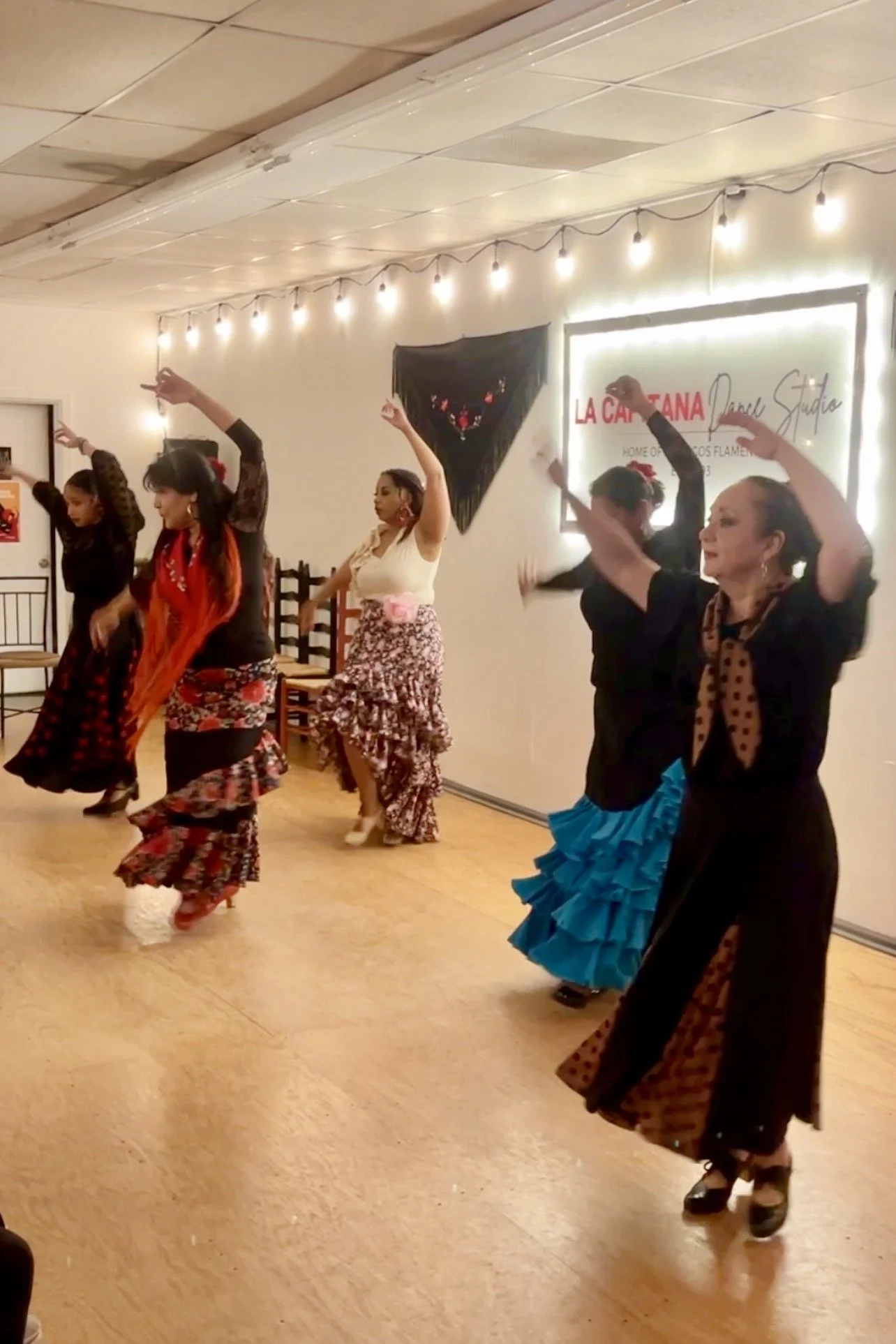 Group of women dressed in traditional flamenco dresses dancing in a dance studio with a sign that reads "La Capitana Dance Studio."