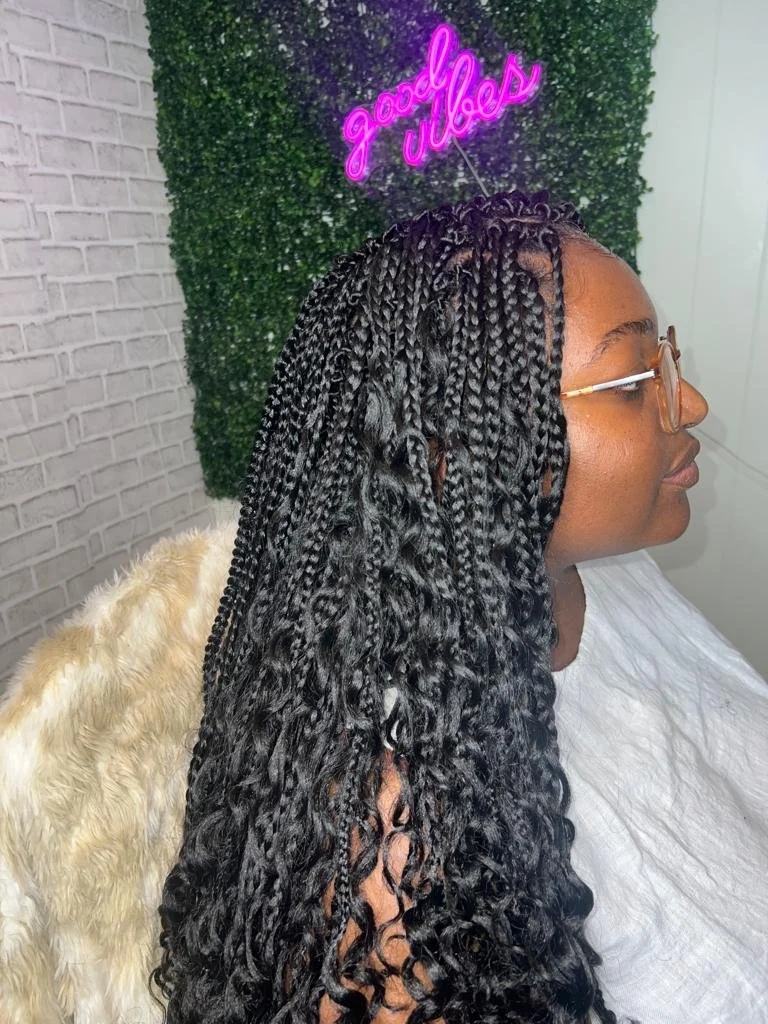 Person with long braids sitting against a faux green wall and pink neon "good vibes" sign.