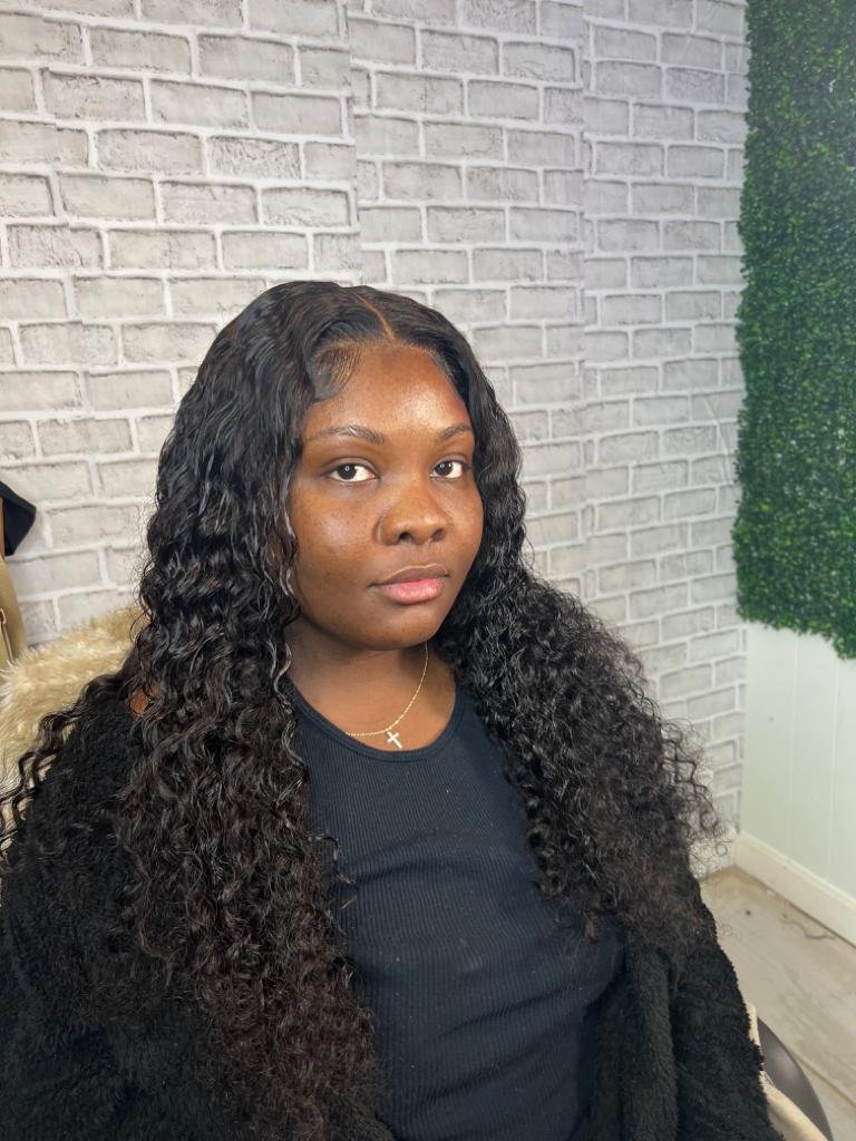 A person with long, curly black hair wearing a black shirt and a gold necklace with a cross pendant, sitting in a room with a light gray brick wall and green foliage on the side.