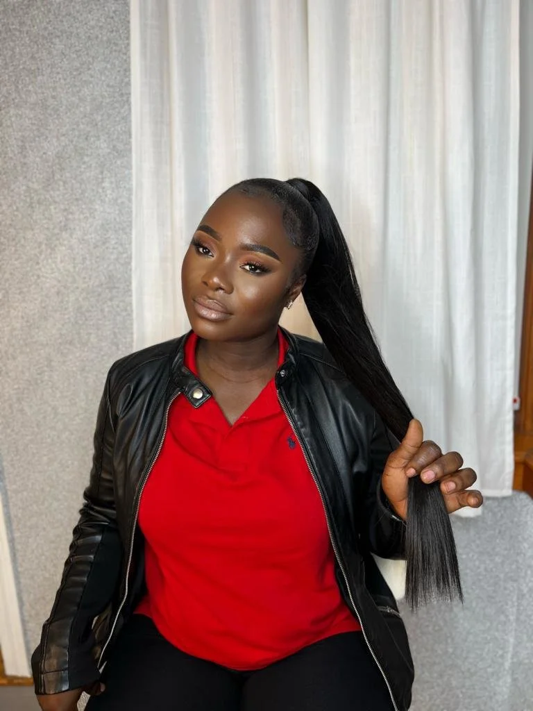 Person with a ponytail, wearing a red shirt and black leather jacket, sitting in front of a curtain.