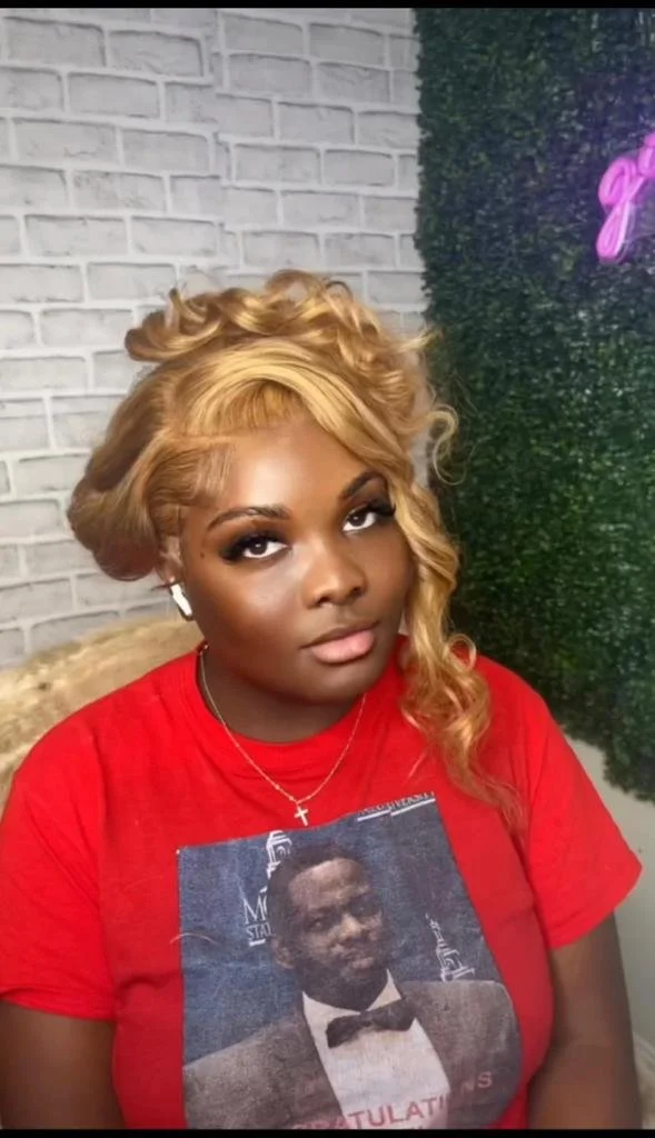 Person with blonde hairstyle wearing a red shirt, featuring a printed image, in front of a brick and foliage background.