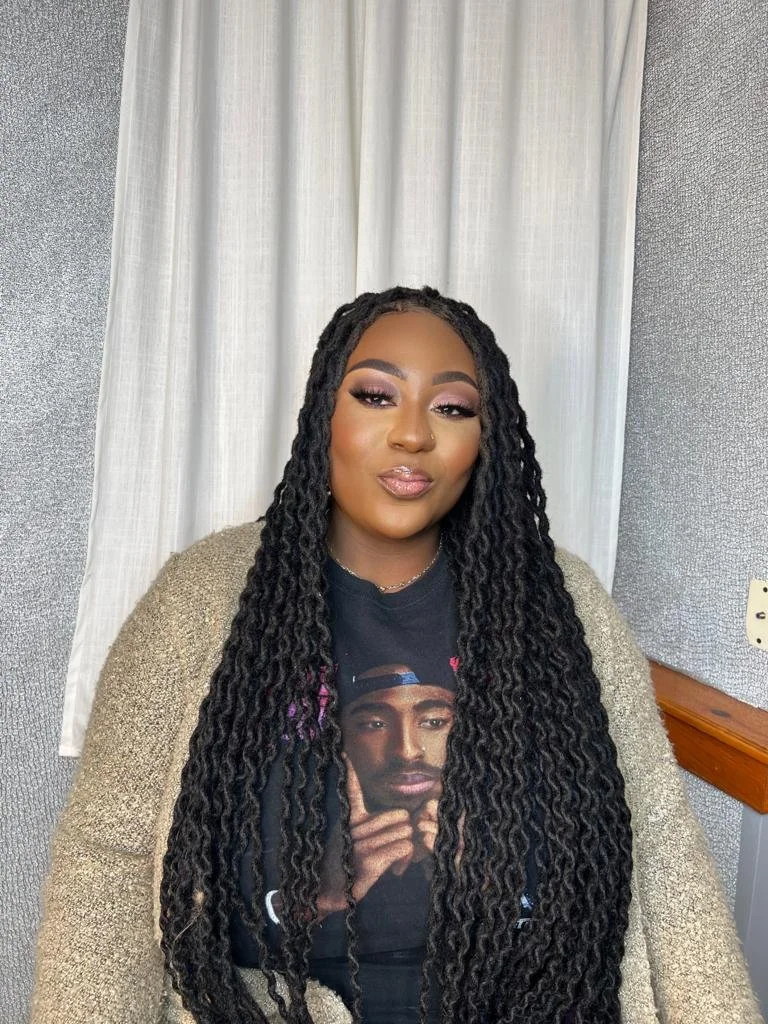 Person with long braided hairstyle, wearing a black T-shirt with a printed graphic, and a beige cardigan, sitting in front of a white curtain.