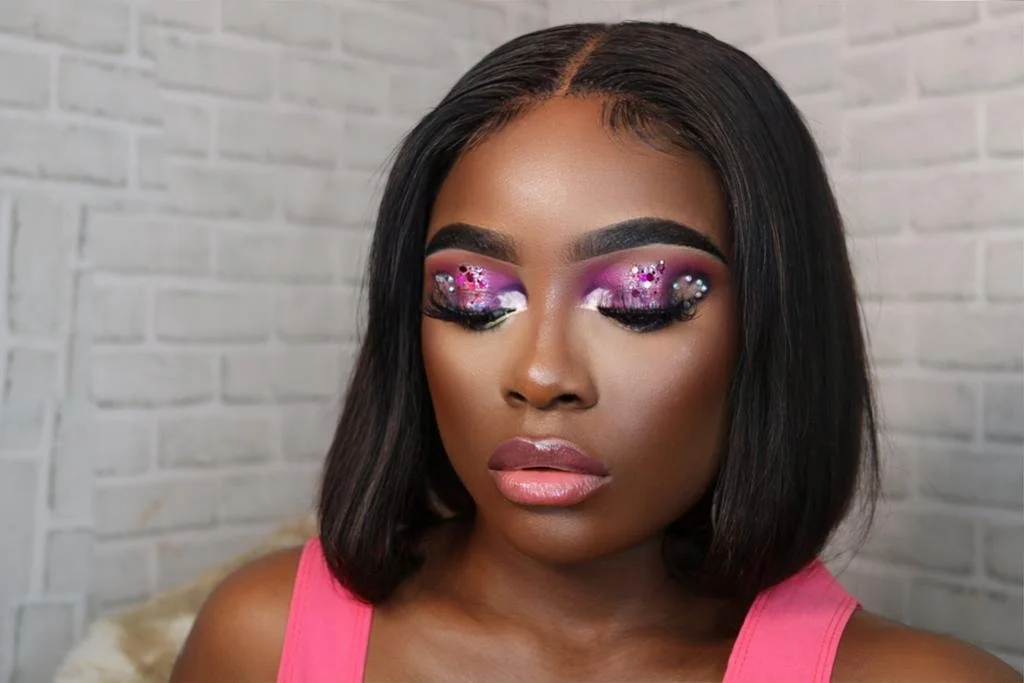 Person wearing colorful eye makeup with rhinestones, pink and purple tones, and glossy lips, against a white brick background.