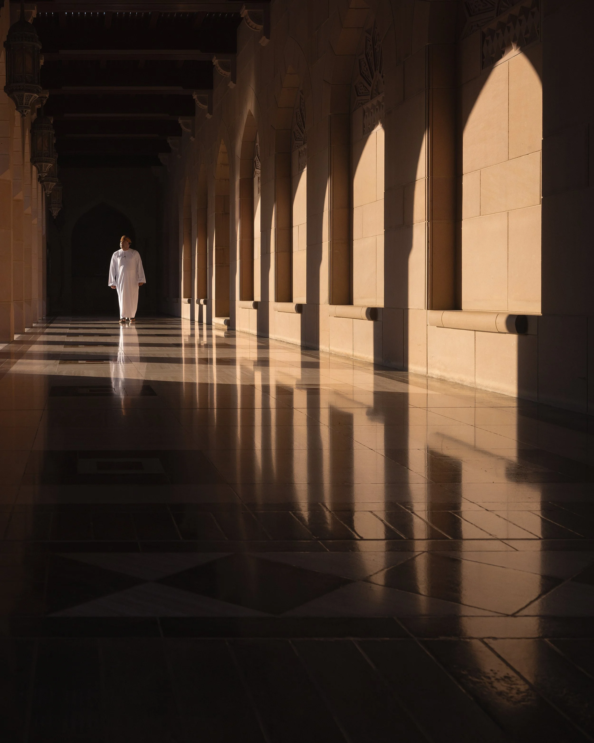 A person dressed in white walking through a corridor with arches at the Sultan Qaboos Grand Mosque in Muscat, sunlight streaming through windows, and reflections on the polished floor.