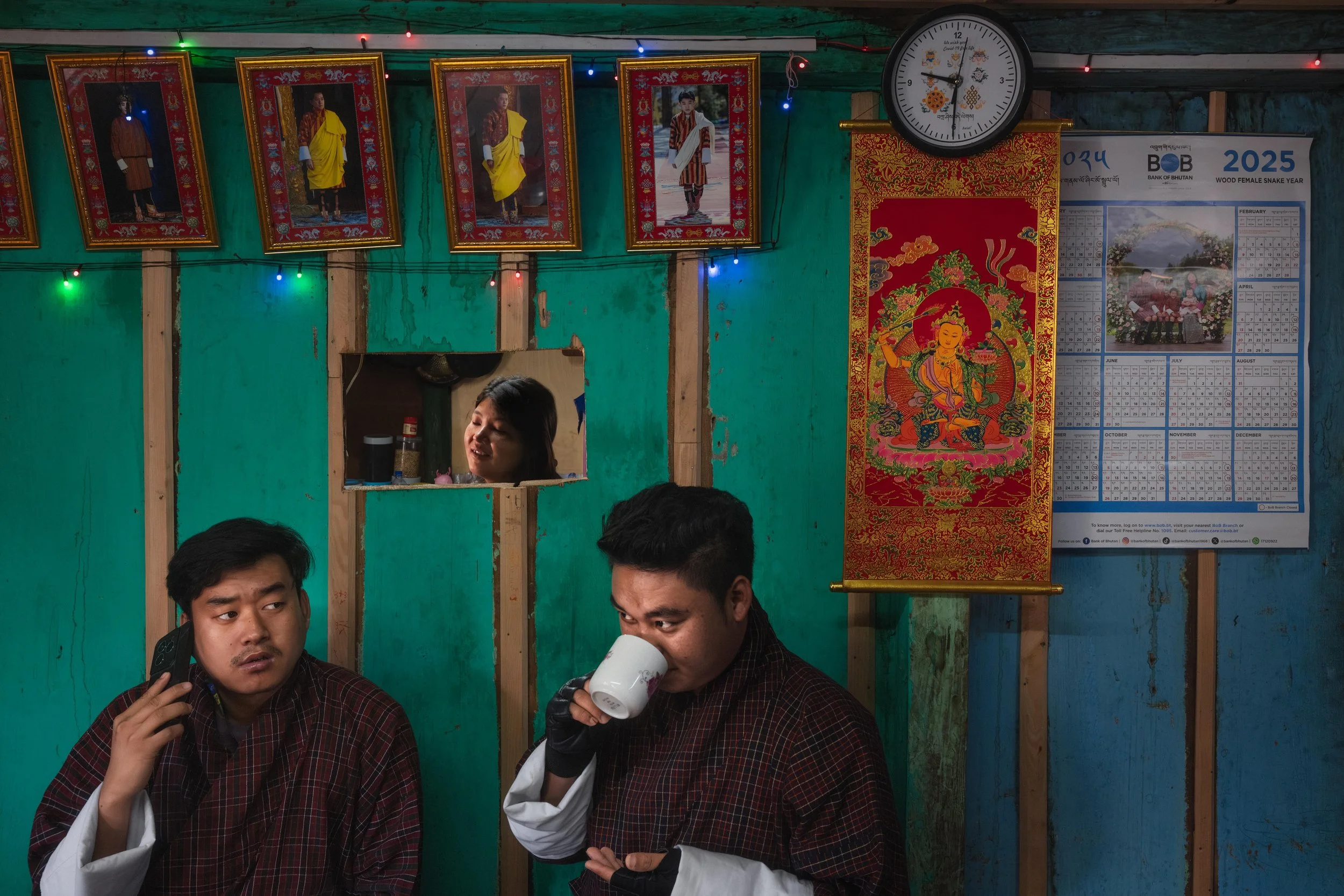 Two young men in traditional Bhutanese attire are indoors. One is talking on a mobile phone, the other is drinking from a mug. The wall behind them is decorated with portraits, colorful string lights, a traditional tapestry, a clock, and a calendar for 2025.