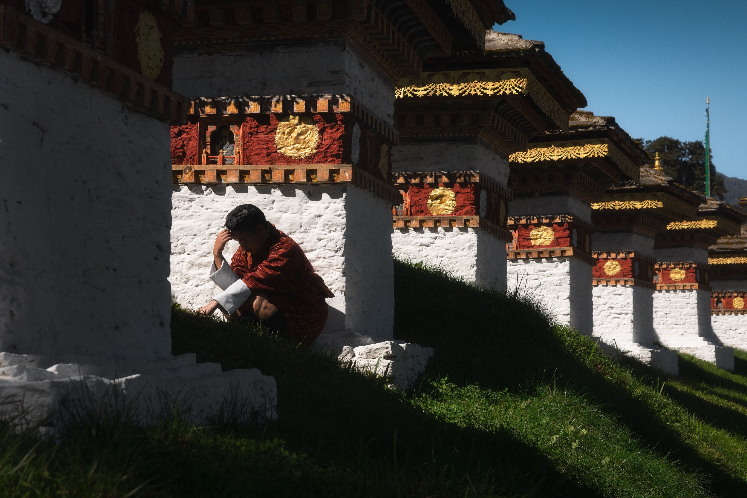 A young person sitting on the grass under a series of white and red ornate Buddhist stupas with golden accents, with a clear blue sky in the background.