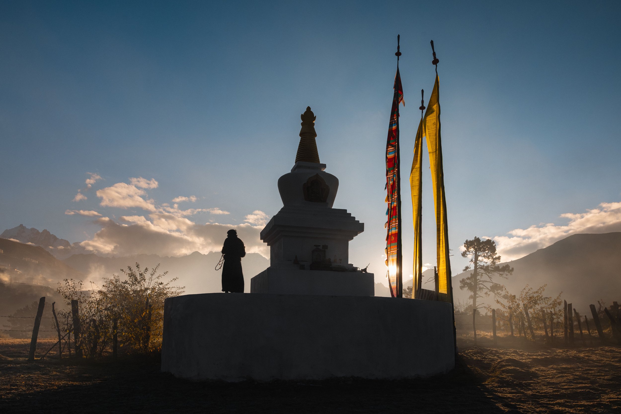 Silhouetted person standing beside a religious stupa with prayer flags flying, mountains and clouds in the background during sunset.