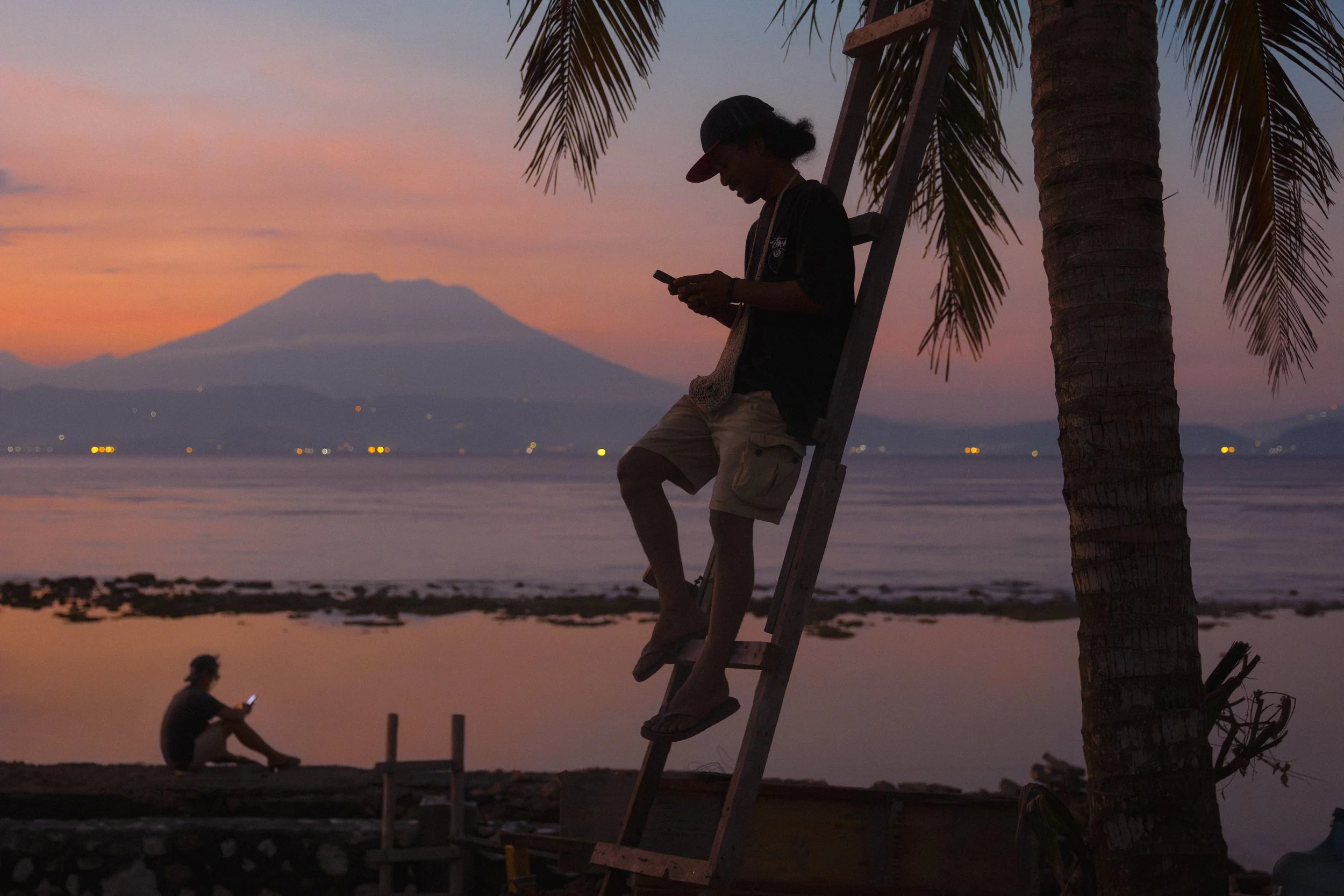 A person wearing a cap and black shirt leaning on a ladder near a palm tree, looking at their phone during sunset, with another person sitting nearby.