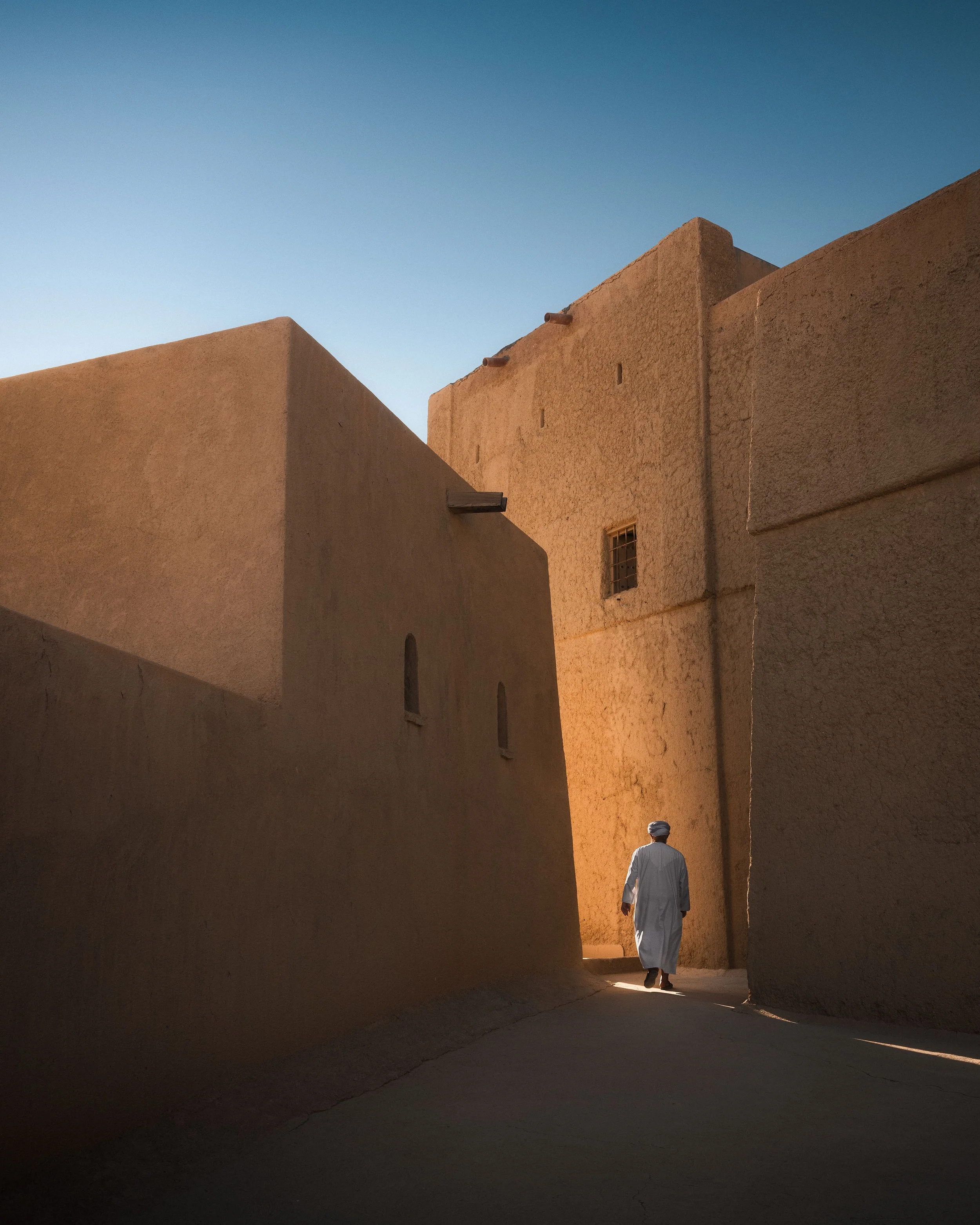 A man in traditional white clothing and a head covering walking through a narrow alley between tall beige adobe buildings under a clear blue sky.