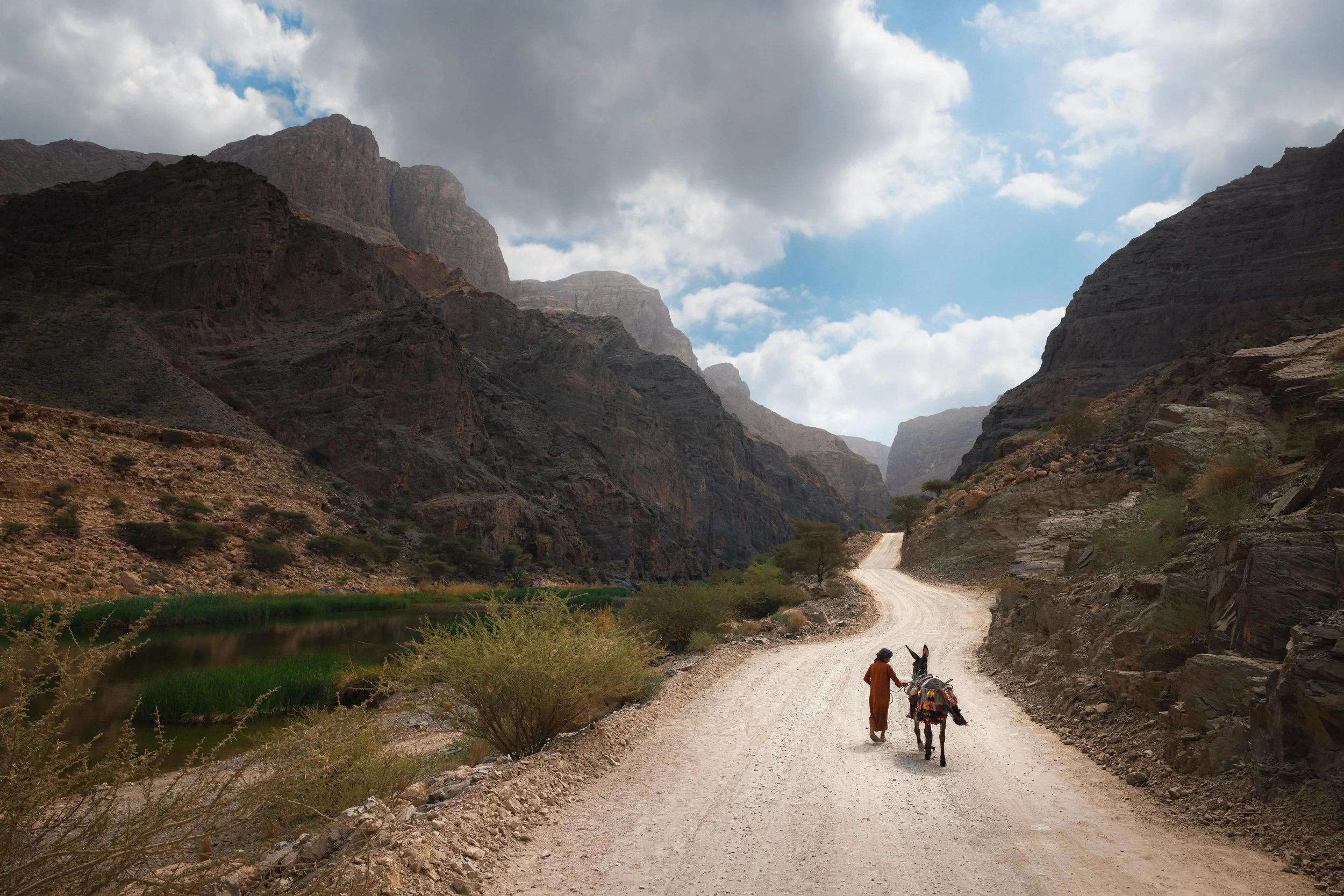 A person walking with a donkey along a dirt road through a mountainous desert landscape, with a river on the left and dramatic clouds overhead.