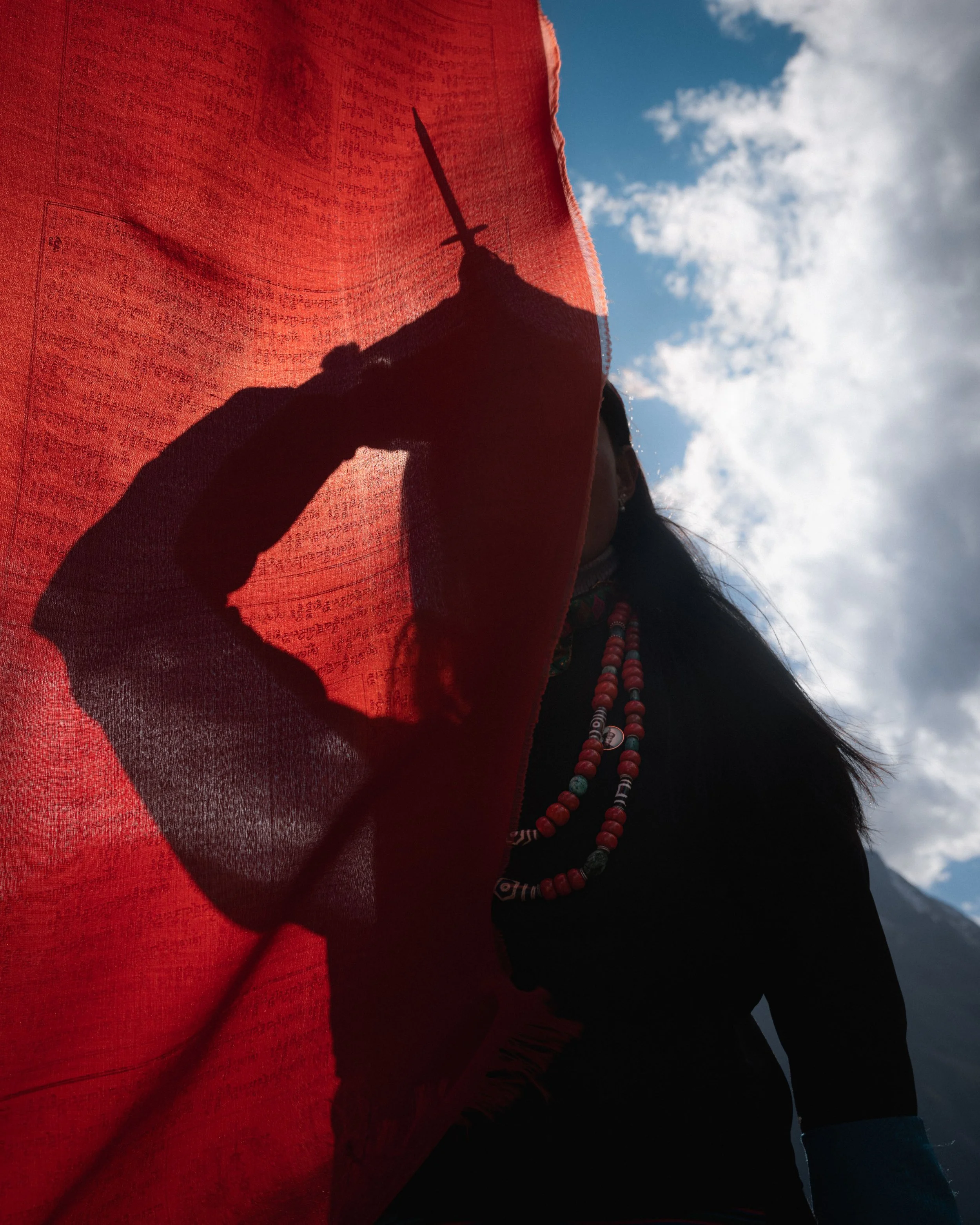 Person holding a red fabric with a shadow of a raised sword silhouette, wearing colorful necklaces against a cloudy sky.