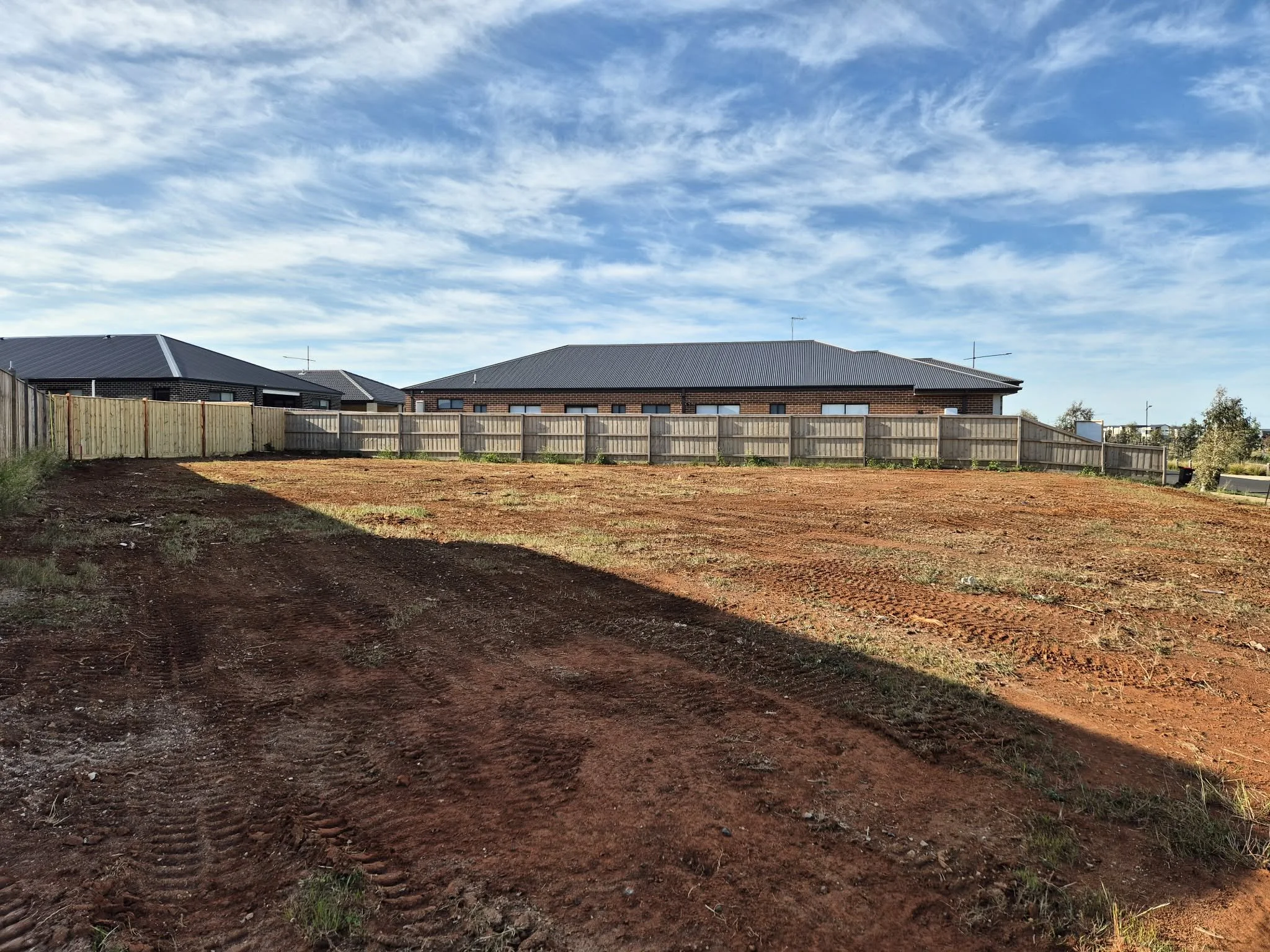 A backyard with a dirt patch, a wooden fence, and modern houses with metal roofs under a partly cloudy blue sky.