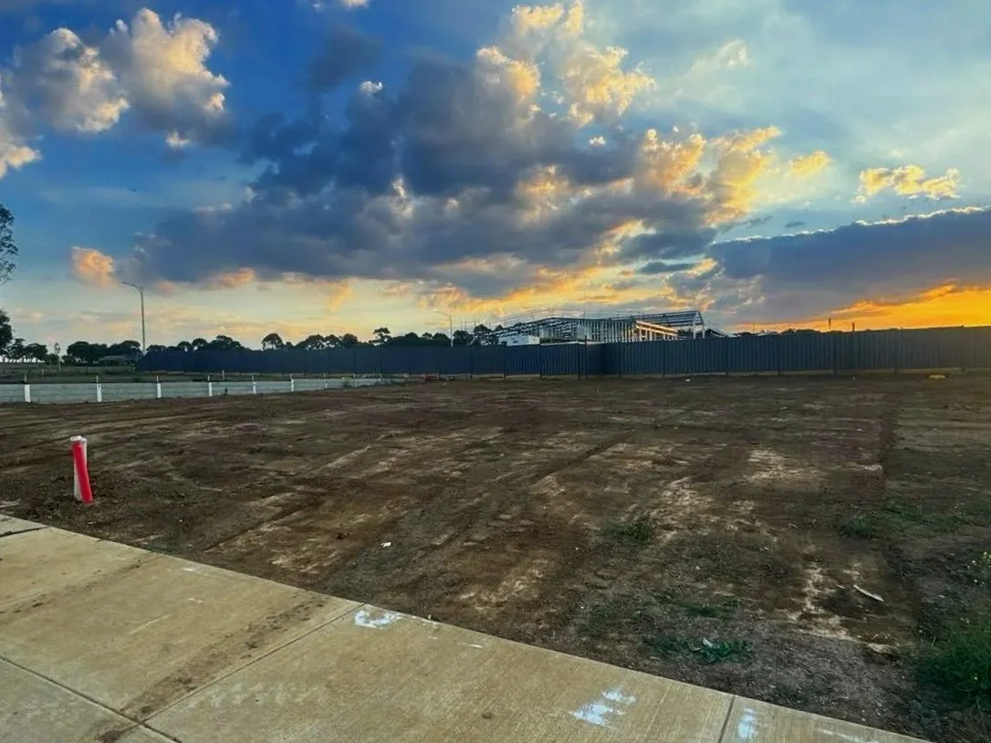 Empty lot with dirt and a sidewalk in the foreground, construction fencing and framework in the background, and a sunset sky with clouds.