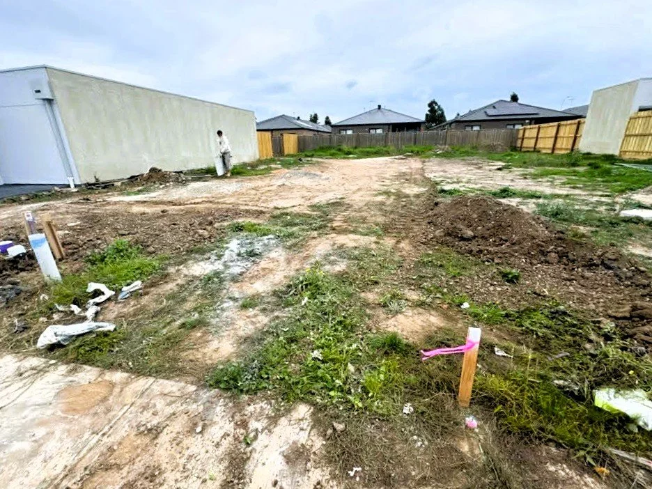 A backyard under construction with dirt piles, a person standing near the side of a house, and houses with fences in the background.