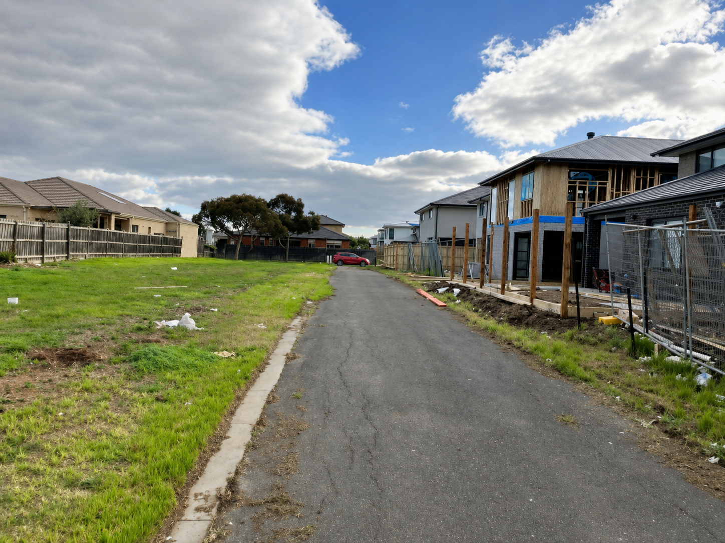 A residential street with a grassy area on the left and a construction site on the right, featuring houses under construction and a cloudy sky.