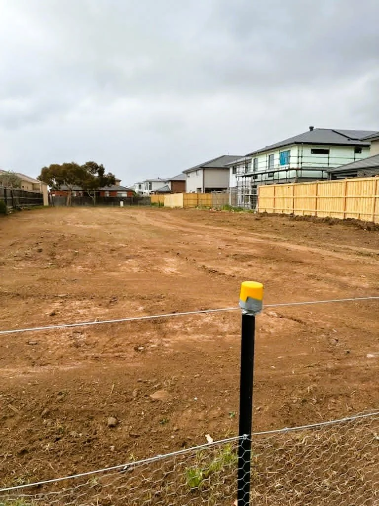 Empty plot of land with dirt ground, surrounded by a fence with houses in the background under an overcast sky.