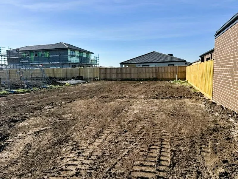 Empty dirt backyard with tire tracks, fenced with wooden fencing, neighboring houses in the background, and a clear blue sky.