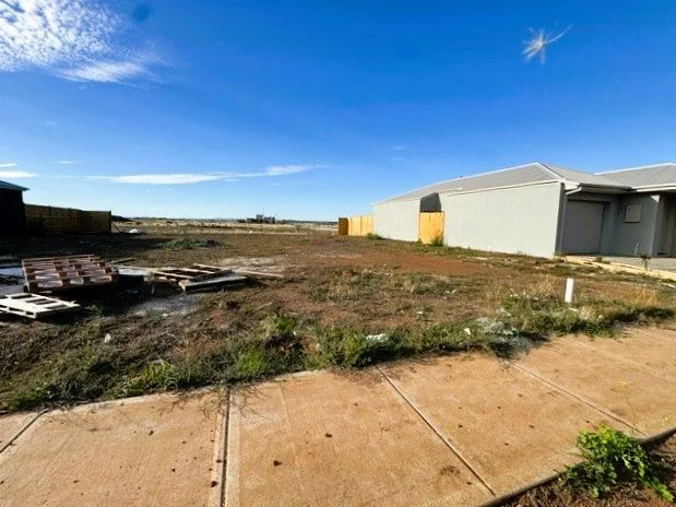 An empty backyard with a concrete patio, sparse grass and weeds, a large metal shed, wooden fences, clear blue sky, and construction pallets on the ground.