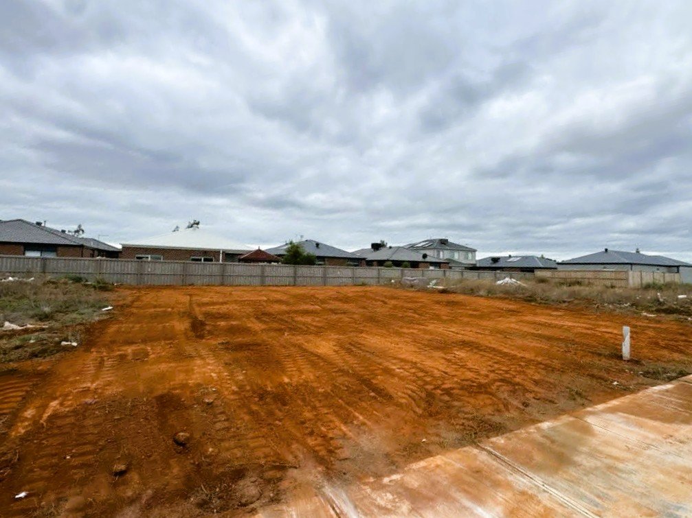 Empty dirt lot with a sidewalk in the foreground and houses with dark roofs in the background under a cloudy sky.