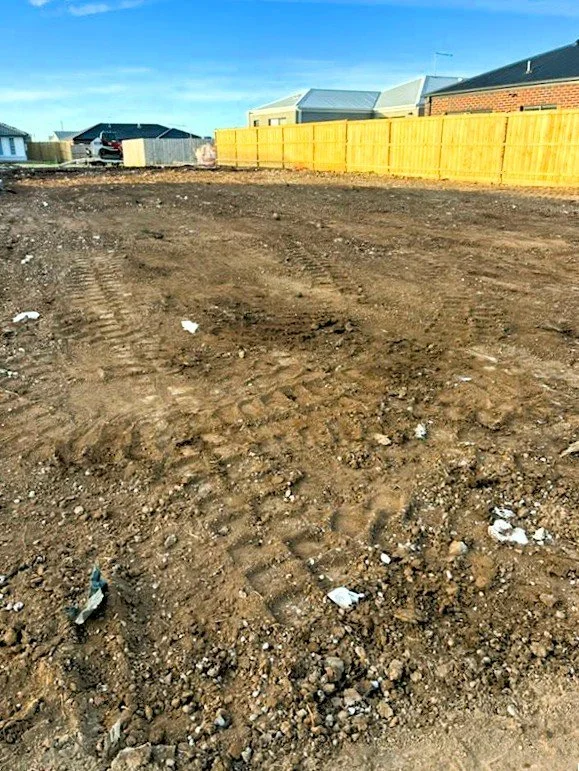 Empty dirt lot with tire tracks, surrounded by houses and a yellow fence, under a clear blue sky.