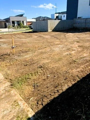 Empty plot of land with disturbed soil and a small wooden stake, in a residential neighborhood with modern houses and a clear blue sky.