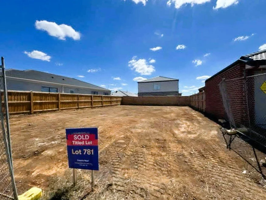 Empty lot with dirt ground, wooden fence, and two houses in the background under a blue sky with clouds.