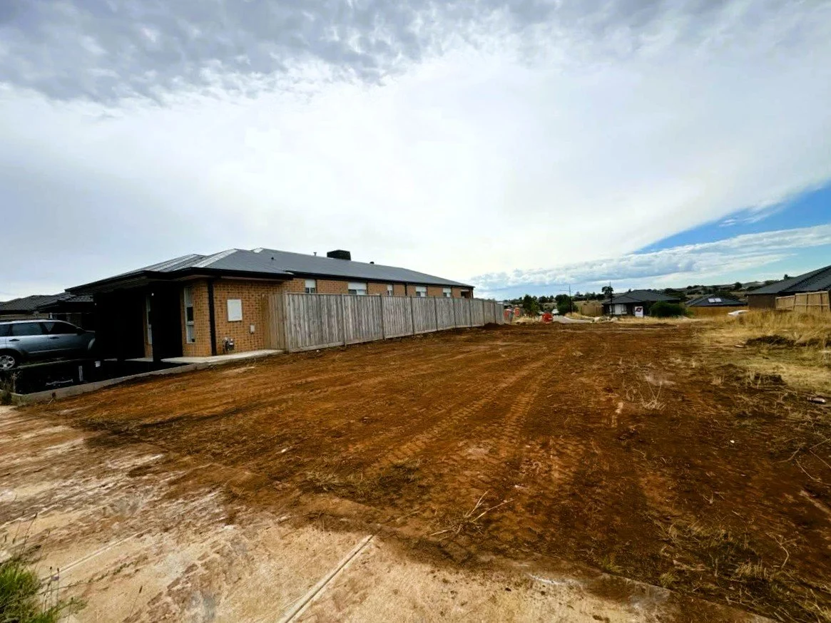 Empty plot of land with freshly cleared soil in a residential area, with houses and parked cars in the background, under a partly cloudy sky.