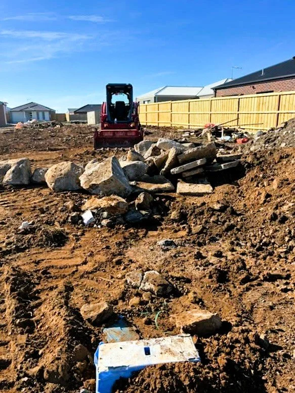 Construction site with dirt and rocks, a small red bulldozer, and a yellow wooden fence in the background.