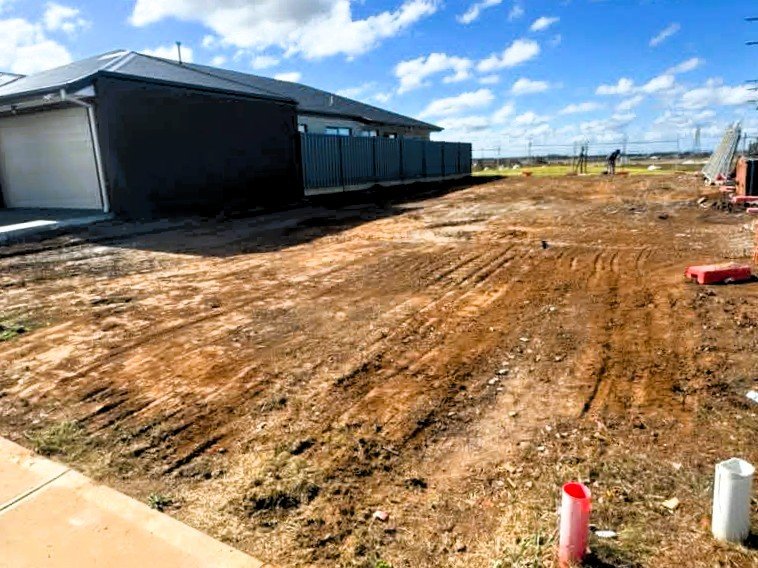 A backyard under construction with dirt ground and partially installed fencing, next to a house with a gray roof and siding, under a blue sky with scattered clouds.