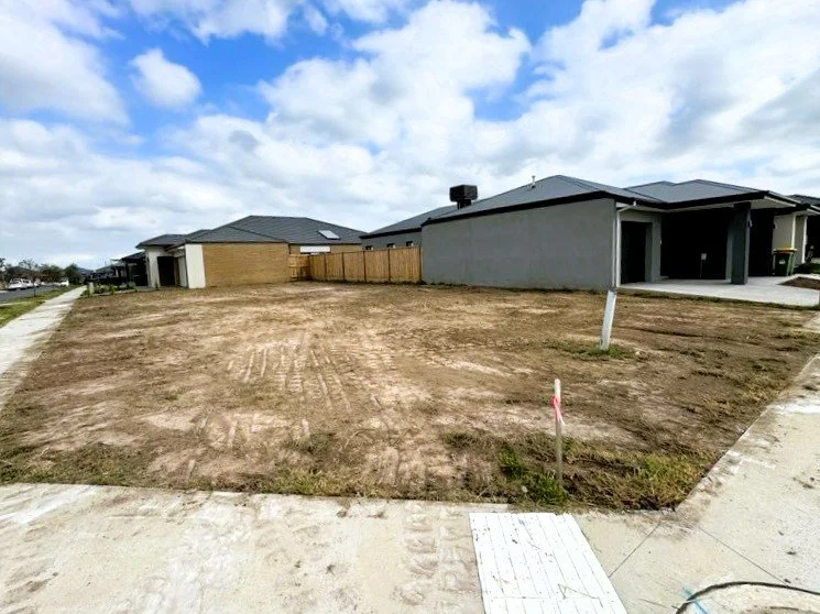 Empty residential lot with dirt surface and adjacent sidewalks, with new houses and fences in the background under a partly cloudy sky.