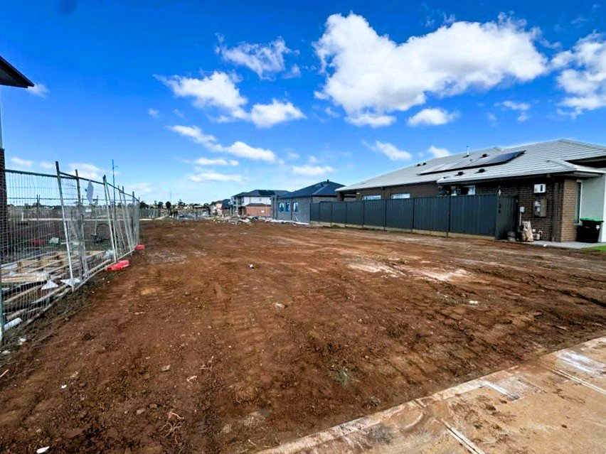 A cleared and leveled dirt lot adjacent to residential houses with construction fencing on the left side under a partly cloudy sky.