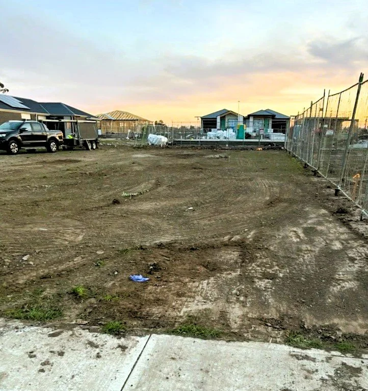 An empty, dirt lot in a residential area at sunset, with construction materials and partially built houses in the background.