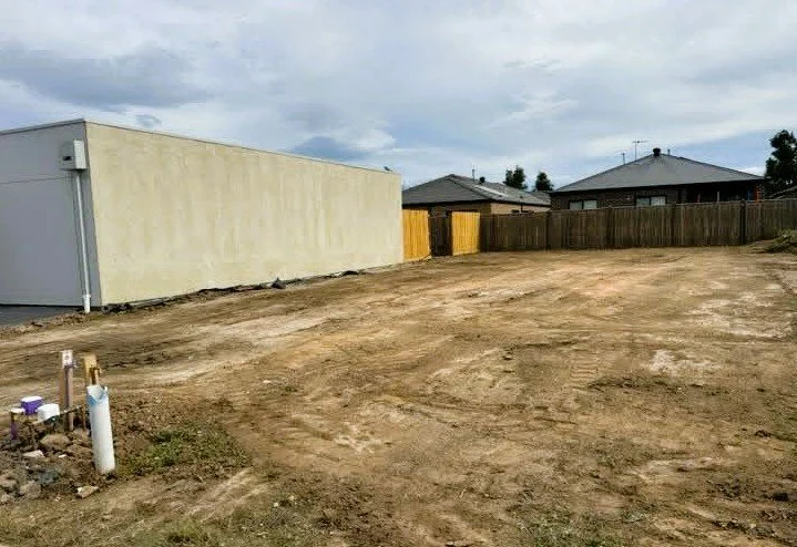 Empty backyard with dirt ground, fenced backyard, and a large beige building with a gray roof.