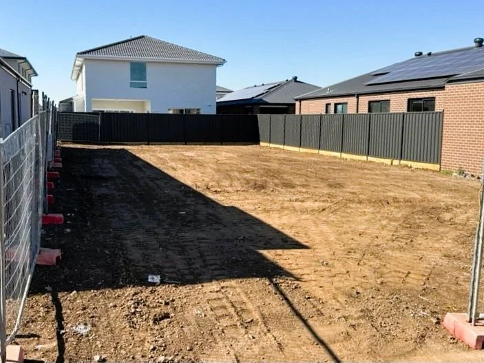 Empty backyard with dirt ground, enclosed by a black metal fence and construction barriers on the sides, with neighboring houses visible in the background under a clear blue sky.