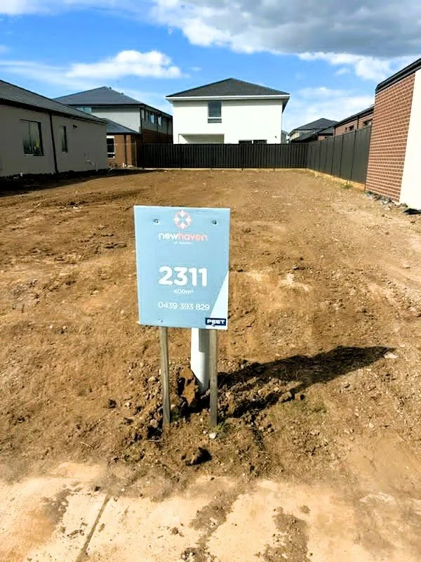 Empty plot of land with a sign that reads "2311" and contains contact information, surrounded by neighboring houses and fencing under a partly cloudy sky.