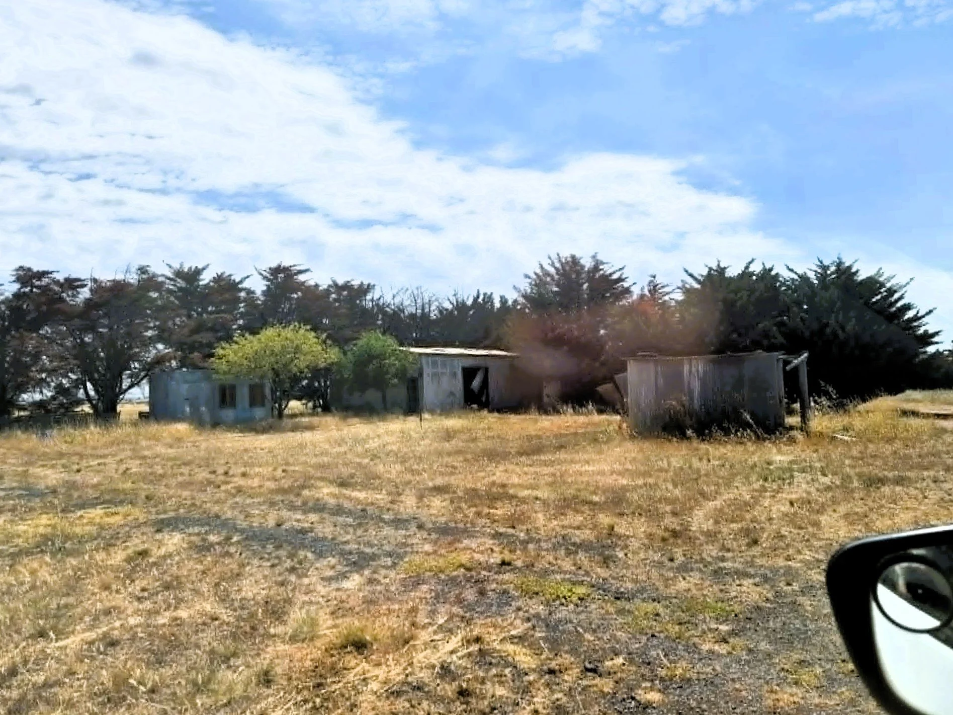 A rural scene with a few old, weathered buildings and trees, a partly cloudy sky, and a car's side mirror visible in the foreground.