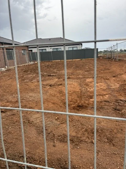 Empty dirt lot behind a wire fence, with houses and cloudy sky in the background.