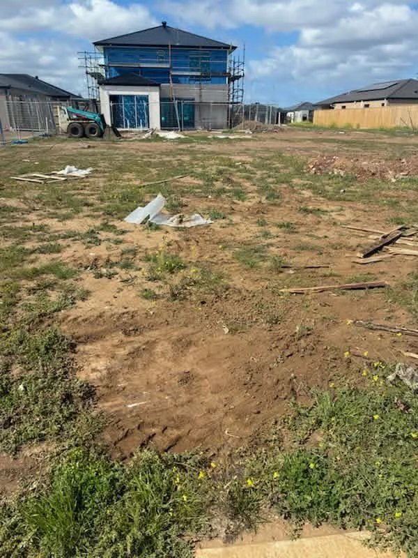A large construction site with a newly built two-story house covered in blue insulation, scaffolding around it, and a cement mixer on the left. The foreground shows dirt and scattered debris, with some green grass and small yellow flowers.
