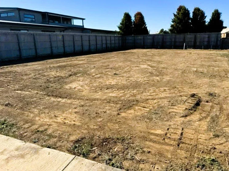 A cleared and leveled dirt lot enclosed by a wooden fence, with a house and trees visible in the background under a clear blue sky.