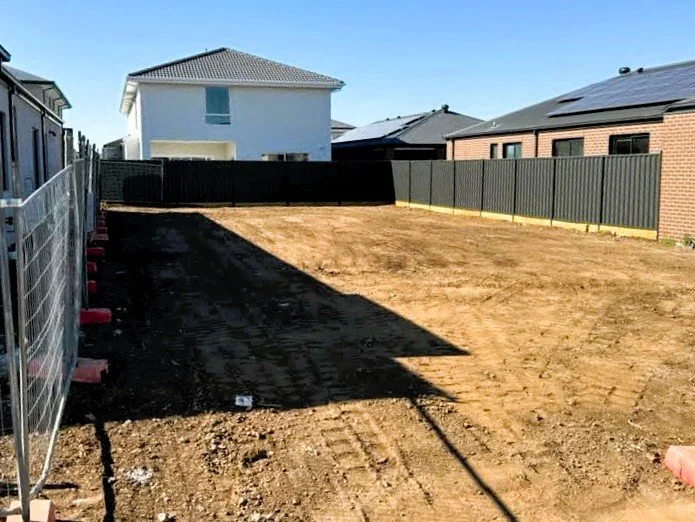 Empty backyard with dirt ground, surrounded by black and gray fences, with neighboring houses visible in the background, under a clear blue sky.