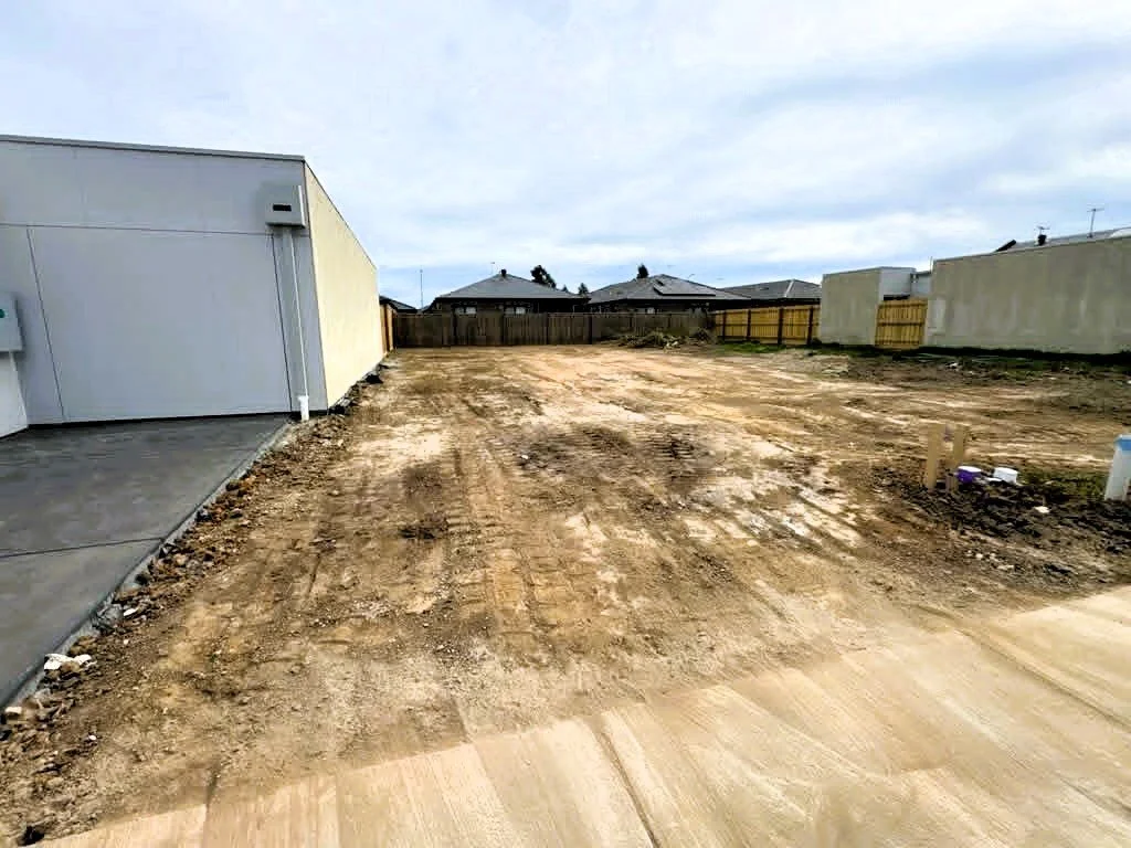 A cleared and leveled dirt lot in a residential neighborhood, with some houses and a fence in the background.