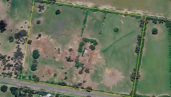 Aerial view of a plot of land outlined by yellow borders, showing grass, trees, and roadside.
